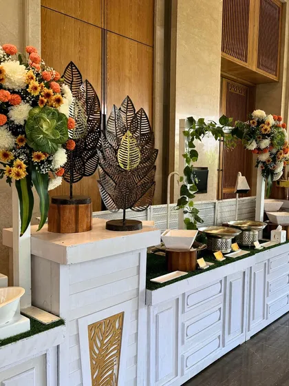 A clean and modern buffet station in white, accented with metal leaf sculptures and lush floral arrangements. This setup demonstrates our versatile design capabilities, suitable for any theme.