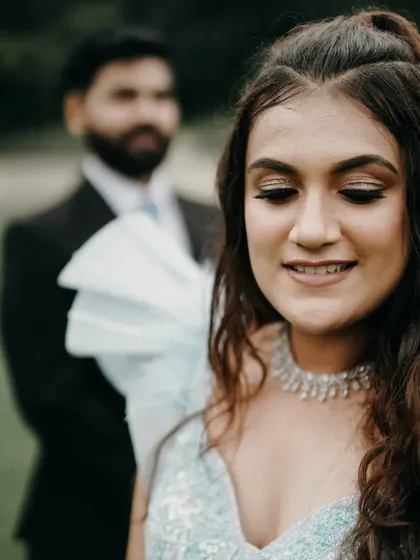 A candid moment from an engagement shoot in a lush garden. The bride is in focus, smiling, while the groom stands slightly behind her, creating a sense of depth and story.
