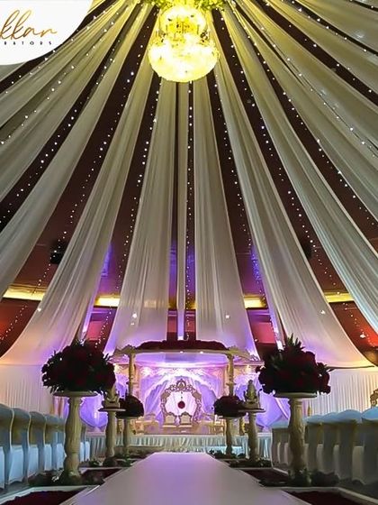 A grand indoor wedding hall with a massive canopy of white drapes and a central chandelier. The aisle is lined with vases of red roses, creating a dramatic and opulent look.