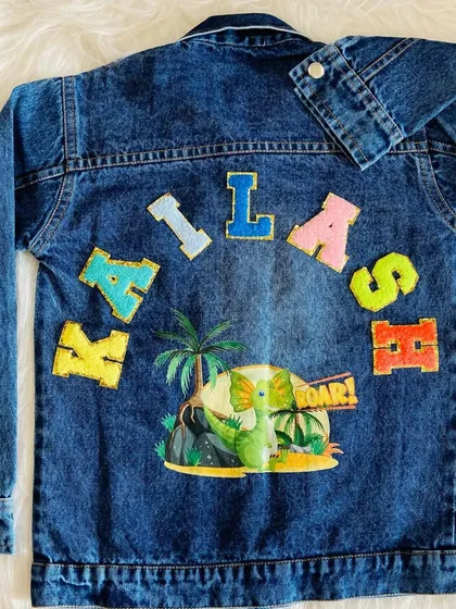 A custom kids denim jacket featuring a dinosaur theme, with the name 'KAILASH' spelled out in colorful patches above a friendly green dinosaur. This shows how we can tailor designs to any child's favorite theme.