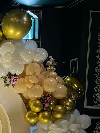 The top corner of the balloon arch, showing a mix of gold, white, and peach balloons with small floral insertions for a touch of softness.
