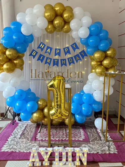 A classic blue, white, and gold first birthday party. The decor features a full balloon ring and a large gold foil number '1'.