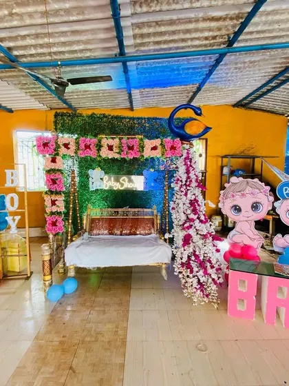A baby shower stage setup in a semi-outdoor space, featuring a decorated swing, a green wall backdrop, and gender-reveal props.