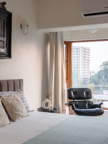 This bedroom blends modern comfort with traditional touches. A classic Eames lounge chair sits by the window, while a vintage carved wooden panel is mounted above the bed as a unique headboard, reflecting the client's personal history.
