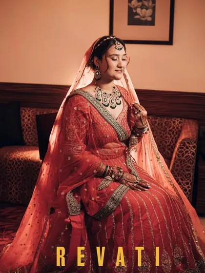 A full-length portrait of the bride, Revati, seated and looking serene in her stunning red lehenga. The soft light beautifully captures the richness of her attire and her calm demeanor.