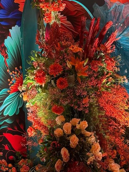 A vibrant and artistic backdrop for a Bhaat ceremony. The design features a colorful, abstract floral wallpaper, complemented by real floral arrangements and whimsical, oversized tulle flowers in shades of red and blue.