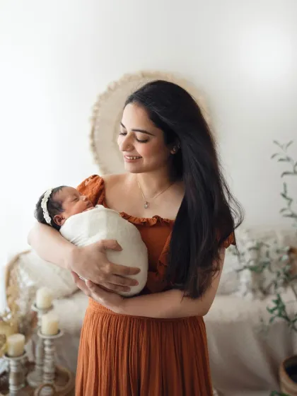 A mother in a lovely rust-colored dress smiles as she holds her sleeping newborn. The bohemian studio setting provides a warm and stylish backdrop.