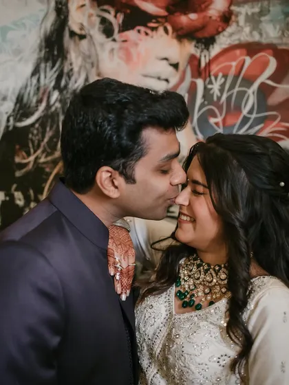 A sweet and candid moment with a gentle kiss on the cheek against a backdrop of modern art. The genuine smile is what makes this engagement photo so special.