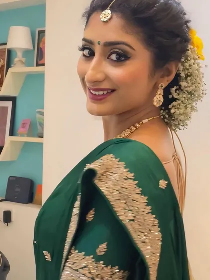 This is a classic engagement look, featuring luminous skin, defined smokey eyes, and a warm lip tone. The hairstyle is an elegant floral bun with soft tendrils framing the face, perfectly complementing the traditional green saree.