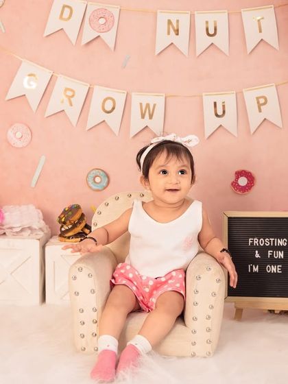 Sitting like a little lady in her chair, surrounded by sweet treats. The "Donut Grow Up" banner adds a playful touch to this first birthday theme.