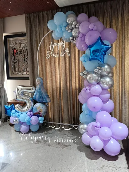 A 'Frozen' themed 5th birthday party setup for a home celebration. The arrangement includes a balloon arch in shades of blue, purple, and silver, with a large '5' foil number and an Elsa character balloon.