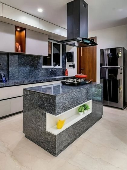 This kitchen features a striking island with a speckled black granite countertop and integrated cooktop. The design is complemented by light-colored cabinets and a glass-fronted display unit with interior lighting.