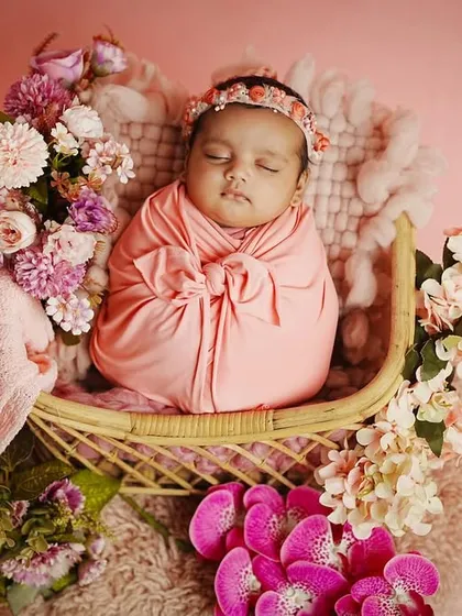A newborn girl sleeps peacefully in a wicker bassinet, surrounded by a cascade of pink and purple flowers. We use a variety of props to create these enchanting setups.