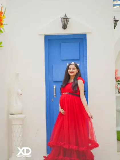 A beautiful solo portrait of the mother-to-be in a striking red gown, standing against a clean white wall with a bright blue door.
