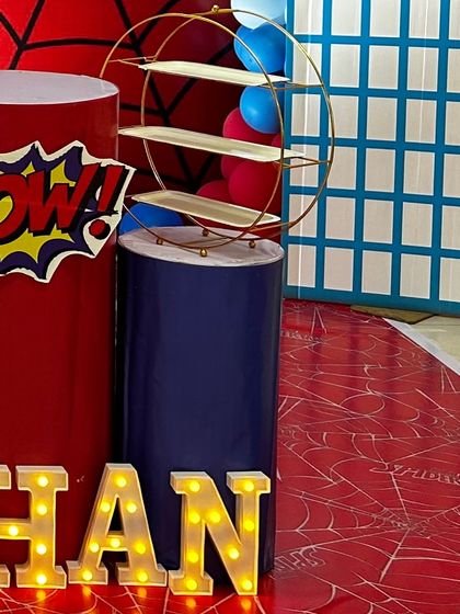 Details from a superhero party, including a "POW!" graphic on a themed plinth and light-up marquee letters. The floor is covered with a spiderweb-patterned mat for a fully immersive experience.