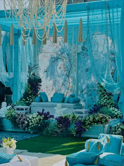 This is my 'Firozi Ishq' theme, a celebration of turquoise. The main seating area features a hand-painted backdrop, framed by flowing turquoise drapes and surrounded by a garden of purple and white flowers.