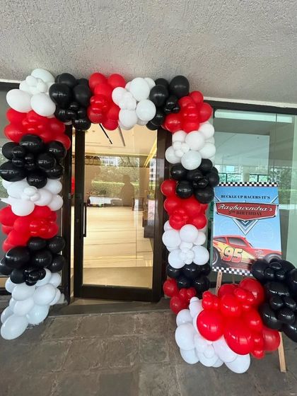 The entrance to a race car party, featuring a balloon arch in red, black, and white. A welcome sign with a Cars theme greets the guests.
