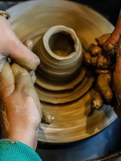 Teamwork at the wheel! An adult's hands guide a child's, showing them the gentle pressure needed to shape the clay. It's a perfect example of learning through guided practice.