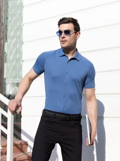 A male model wearing a steel blue short-sleeved shirt and black trousers, posing confidently outdoors. The bright, natural lighting highlights the shirt's color and athletic fit, suitable for a brand's social media or website.