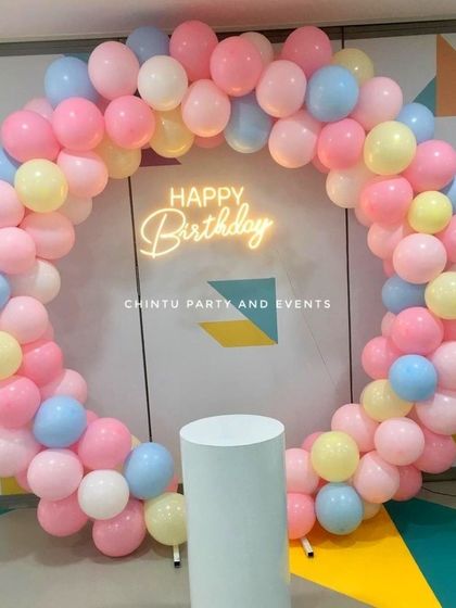 A simple and sweet pastel balloon ring for a birthday at home. The mix of pink, yellow, blue, and white balloons creates a cheerful backdrop, complete with a neon sign and a pedestal for the cake.