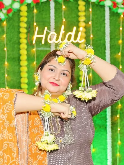 A happy client at her Haldi ceremony, wearing a bright yellow floral jewellery set. The set includes a maang tikka, earrings, and haath phool with delicate hanging flowers.