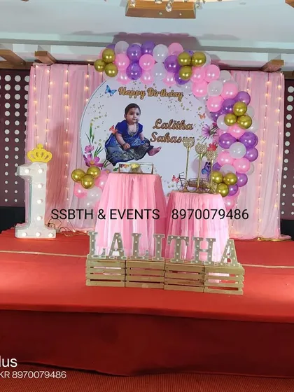 A princess-themed first birthday on a stage. The decor features a pink and purple balloon arch, a personalized backdrop, and a light-up number '1' with a crown.