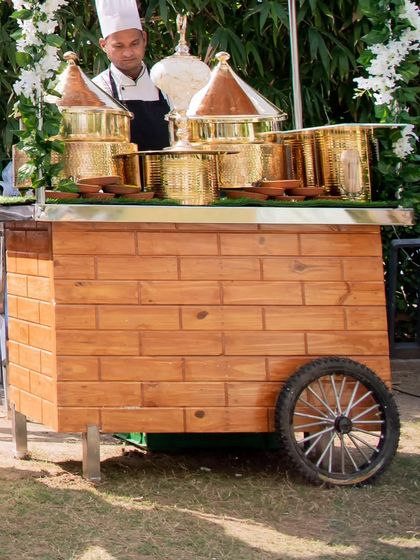 A close-up of a rustic wooden food cart adorned with ornate brass serving vessels. This setup combines traditional Indian aesthetics with a professional catering service.