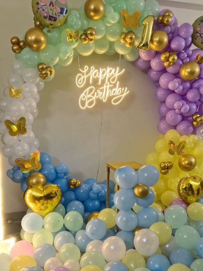 A dreamy pastel-colored first birthday setup. The balloon ring is made of light blue, yellow, and purple balloons, with gold accents, a neon sign, and a large marquee number '1'.