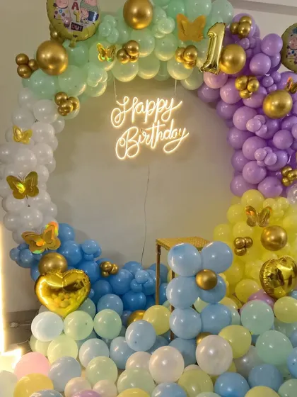 A bright and cheerful first birthday decoration using a full circle balloon arch. The pastel rainbow colors, 'Happy Birthday' neon sign, butterfly accents, and a large light-up '1' make this a perfect spot for birthday photos.