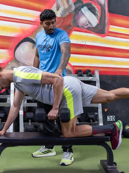 A coach provides tactile cues to an athlete during a single-arm dumbbell row on a bench. This exercise targets unilateral back strength and core stability, essential for rotational athletes and overall postural health.