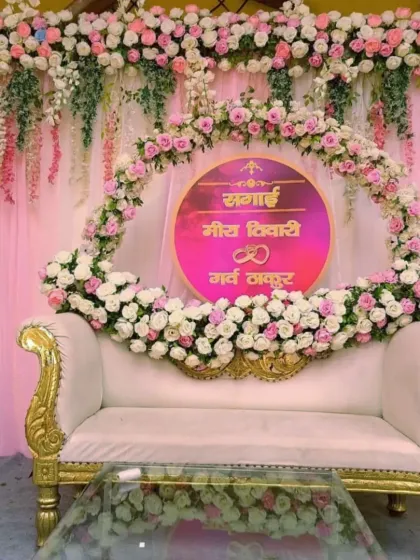 A beautiful and personalized stage for a Sagai ceremony, featuring a floral ring with the couple's names, a plush white sofa, and a backdrop of pink drapes and flowers.