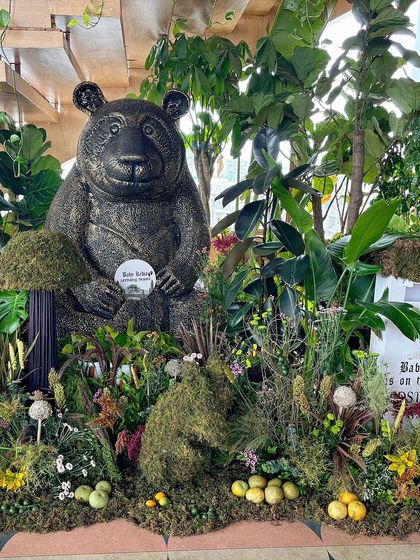 A whimsical baby shower setup featuring a large bear statue, a 'post here' mailbox for wishes, and a lush forest floor of moss and flowers.