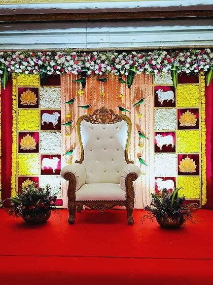 A traditional backdrop for a Seemantham ceremony featuring a checkerboard pattern of flowers and fabric. The design includes auspicious elements like cow motifs and lotus cutouts, set against a rich red curtain.
