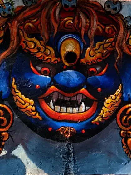 A painting of a blue deity mask with a third eye, rendered with fiery red and gold details. This work draws from mythological iconography to create a powerful and intense visual statement.