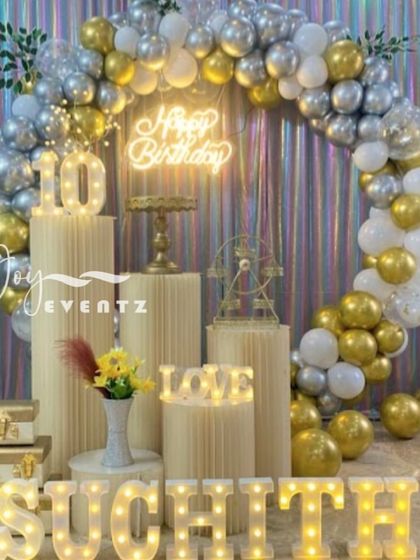 Here is a sophisticated ring balloon decoration I did for a 10th birthday. The setup includes a full arch of metallic gold, silver, and white balloons, a holographic curtain backdrop, an illuminated number '10', and marquee letters spelling the name 'SUCHITH'.