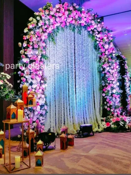 An enchanting floral archway perfect for a wedding reception or engagement. We crafted this stunning entrance using a cascade of white jasmine flowers framed by a lush arch of pink and purple roses, accented with warm lanterns for a romantic glow.