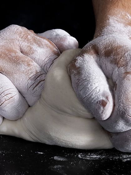 For our pizzas, we prepare a different kind of dough. Here, hands dusted with flour work the dough to create the perfect base for our wood-fired pizzas.