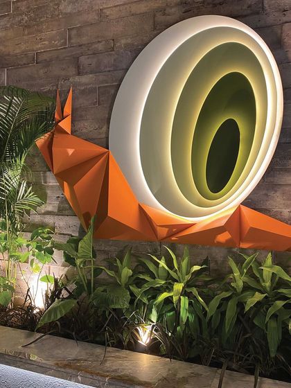 The illuminated snail art, set against a textured stone wall and surrounded by tropical plants, demonstrates our passion for integrating biophilic art and lighting.