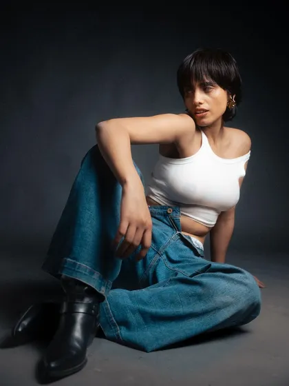 A relaxed, seated pose in a studio setting. The lighting highlights the textures of the denim and the ribbed tank top, creating a casual yet polished editorial look.