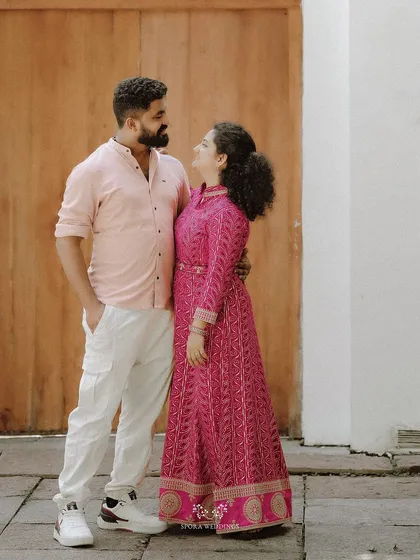 A full-length portrait of the couple, her in a vibrant pink dress and him in a matching pink shirt, looking lovingly at each other.