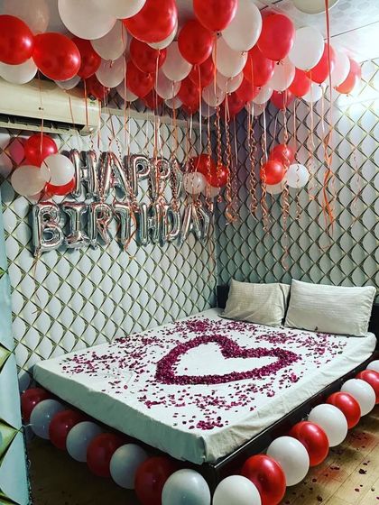 A beautiful birthday room setup with a red and white color scheme. The decor features 'Happy Birthday' foil balloons against a textured wall, a heart of rose petals on the bed, and balloons lining the base of the bed.