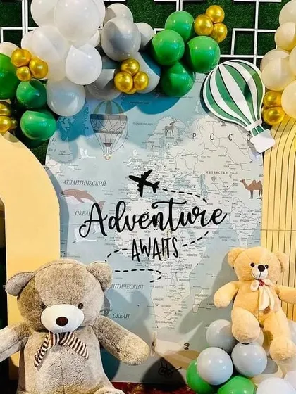 Detail shot of the "Adventure Awaits" backdrop. The design features a world map, airplane graphics, and cute teddy bears, all framed by green, white, and gold balloons.