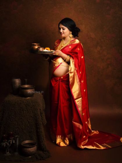 The expecting mother holds a traditional thali (plate) with offerings, a beautiful symbolic gesture in this culturally rich maternity portrait. Her red saree and exposed baby bump are the focus of the shot.