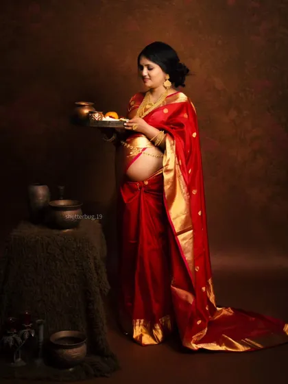 The expecting mother holds a traditional thali (plate) with offerings, a beautiful symbolic gesture in this culturally rich maternity portrait. Her red saree and exposed baby bump are the focus of the shot.