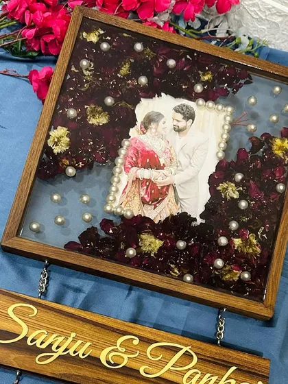 A 12-inch square teakwood frame with a hanging nameplate. The frame preserves red wedding garlands around a central photo.
