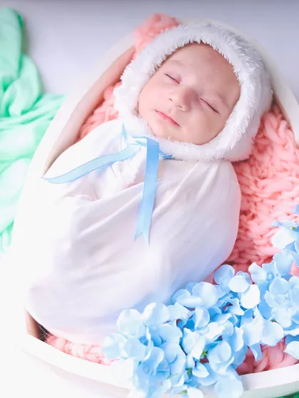 A newborn dressed as a little bunny with a fluffy white bonnet, sleeping in a heart-shaped bowl. The soft pink and blue colors create a gentle, pastel palette.