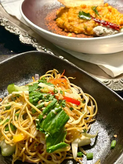 A pairing of our global culinary offerings. In the foreground, a vibrant Asian noodle salad, and in the background, a contemporary take on a South Indian dish, showcasing our ability to plate diverse cuisines with precision and style.