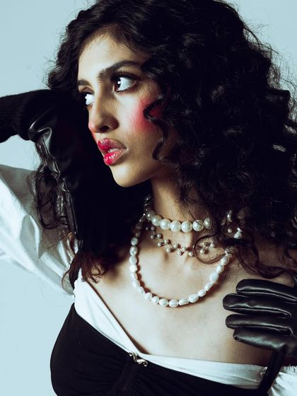 A close-up on the character's face. The heavy blush, glossy lips, and wide-eyed look are all part of the theatrical styling. The hands in black gloves add a touch of drama and contrast.