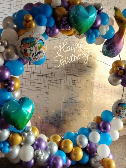 A mermaid-themed balloon ring in a mix of blue, purple, gold, and silver. The design features a mermaid tail, heart-shaped foil balloons, and a "Happy Birthday" neon sign.