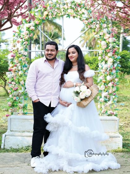 A romantic couple's portrait in front of a floral arch. The mother-to-be is wearing an angelic white ruffled gown, perfect for a dreamy maternity shoot.